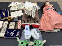 Full lot overview showing bisque doll, knitted slippers, jewelry pieces, handkerchiefs, and small decor items arranged on table.