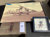 Photo showing a wooden box, a square baseball-themed clock with a baseball pitcher and baseball hour markers, and a large photo art print of a Navy helicopter.