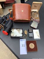 Full lot showing binoculars, leather case, and box of jewelry items on table with auction number card.