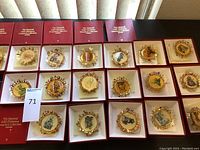 Wide shot displaying 20 gold Hummel Christmas ornaments in red boxes, arranged in four rows of five. Each ornament features a round illustration of Hummel characters with ornate gold metal edges.