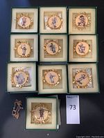 Ten Hummel gold Christmas ornaments in green cushioned boxes arranged in a grid. Each ornament has a gold-tone frame with detailed festive charms and central Hummel figurine illustration. A metal necklace chain is also present beside one box.
