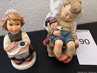 Two Hummel Erste Ausgabe special edition porcelain statues on black surface, girl holding tray and boy with doll visible