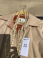 Close-up of beige London Fog trench coat hanging on hanger with a white floral necklace placed on it and lot number 21 tag attached