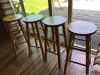 Four wooden oak bar stools placed in a row by a window. One stool has paint marks on one leg visible.
