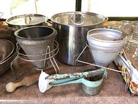 Photo showing conical sieves on stands, metal pots, and a vintage apple peeler along with a wooden pestle on countertop