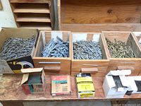 Photo showing four wooden boxes filled with assorted nails and several boxes of staples placed in front on a wooden surface.