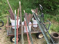 Overview of lot showing metal fence posts, rebar in buckets, wire spool, and metal utility wagon in grassy outdoor area.