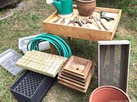 Overview of gardening potting table with hand tools arranged on top, green watering can, coiled garden hoses, several planting trays and compost sifter, and stack of square wooden planter bases with round flower pot on grass