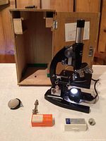 Microscope setup with light source lit, wooden carrying case open behind, and microscope accessories displayed on table.