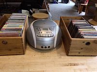 Presidian silver portable CD player with cassette deck placed between two wooden crates filled with CDs.