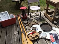 Photo shows the cooler, kerosene heater, wooden oars, cast iron cookware, tackle bag and small outdoor items all arranged on weathered wooden deck surface.