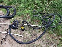 Wide view of two sump pumps connected to black corrugated hose lying on soil and grass.