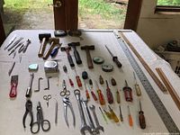 Wide overview of assorted small hand tools including hammers, screwdrivers, pliers, scissors, rulers, and measuring tapes spread on table.