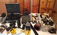 Overview of various cameras, lenses, battery chargers, and accessories on a table with a wood panel wall in background.