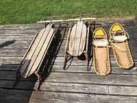 Two wooden sleds and one pair of snowshoes laid out on a wooden deck in sunlight.