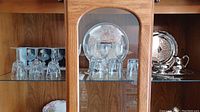 Display cabinet with crystal stemware, glass tumblers, and silver plated serving trays and teapot arranged