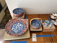 Shows entire lot with three bowls and two cups situated on wooden boxes for display. Exhibits detailed patterning, color combination, and visible imperfections.