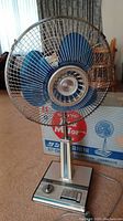 Full view of vintage Sanyo fan with blue blades and protective metal grill, standing on carpeted floor with original box in background.