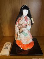 Front view of Japanese doll showing detailed kimono, black hair and porcelain face with closed eyes, standing on black rectangular base.