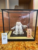 Full view of Japanese doll inside a black framed glass display case on an orange carpet background. A lot tag and wooden plaque are visible inside the case.