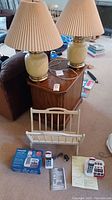 Pair of brass and porcelain table lamps with beige pleated shades standing on wooden table alongside folding wooden magazine rack, cordless phones and boxes on carpeted floor