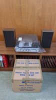 Full view of turntable on shelf in plastic wrap, two speakers set aside and original cardboard box beneath.