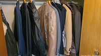 Photo of multiple men's suits and sports coats hung closely together on a closet rod, showing different colors and styles including dark blue pinstripe, tan, and dark patterned fabrics.