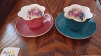 Two Crown Staffordshire teacups on matching saucers, one teal and one maroon, showing scalloped edges and fruit design inside cups.