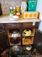 Wide view of vintage kitchen items arranged on countertop and inside open cabinet, showing pots, pans, thermos, cups, plates, and storage containers