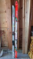 Two pairs of skis standing vertically, one bright orange Atomic ASC Kevlar System 80 with white and green lettering, the other gray TM Elan skis with purple and pink design, both showing scratches.