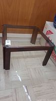 Square wood framed side table with clear removable glass top shown in room with wood paneling background.