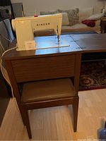 Singer sewing machine mounted in wooden cabinet with matching stool underneath. Machine appears cream colored with Singer branding.