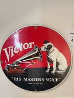 Round Victor "His Master's Voice" hanging plaque with dog and gramophone design, red, black, and white enamel, new in shrink wrap.
