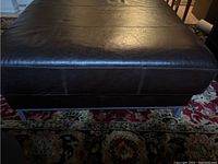 Front side view of modern black leather ottoman showing stitch detail and chrome legs on a patterned rug.