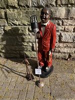 Full view of two sculptures: the carved jazz singer in red suit with old-fashioned standing microphone and the small metal trumpeter sculpture beside it, positioned outdoors on a paved ground with stone wall background.