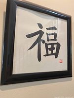 Framed artwork featuring a black Japanese character on a white background in a black frame.