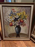 Framed painting showing a blue vase filled with a variety of flowers including daisies and yellow blossoms on a round wooden table.