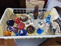 Photo showing an assortment of glass paperweights, figurines, and a ceramic cup and saucer set inside a white basket on yellow fabric.