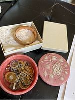 Pink round container with lid holding assorted mixed vintage jewelry including bangle, necklaces, earrings, pendants, and rings.
