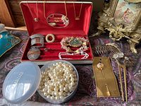 Overview of assorted jewelry laid out on a fabric surface, including pearl necklace in a container, bracelets, pins, and earrings in a red jewelry box.