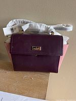 Front view of Kate Spade handbag showing purple flap and pink sides with handles wrapped in protective material.