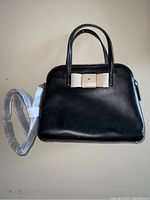 Full view of the black handbag with beige bow and white shoulder strap wrapped in protective plastic.