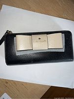 Front view of the black leather wallet showing the beige leather bow detail with gold embossed Kate Spade New York logo.
