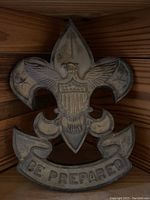Metal crest shaped like a fleur-de-lis with eagle shield emblem and 'Be Prepared' banner, showing detail and wear