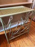 Full view of mint green metal table showing woven pattern top and decorative leaf and white flower metalwork on the sides.