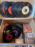 Box containing multiple 7-inch vinyl 45 RPM records stacked and leaning against the side of the box, alongside audio cassette tapes in cases with cover art visible