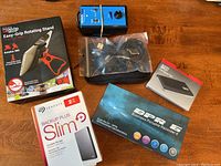 Photo displaying Aduro uGrip Easy-Grip Rotating Stand box, a blue/black device with switches, sealed bag with cables, DPR recorder box, and Seagate external hard drive box on wooden surface.