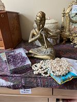 Photo showing brass Buddha statue playing a flute with purple bead necklace and faux pearl necklace on a patterned cloth