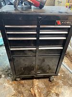 Front view of Excel black metal tool cabinet with multiple drawers and lower doors, showing use wear and dirt on casters and surfaces.