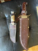 Two vintage machetes side by side with their brown leather sheaths on a black surface.